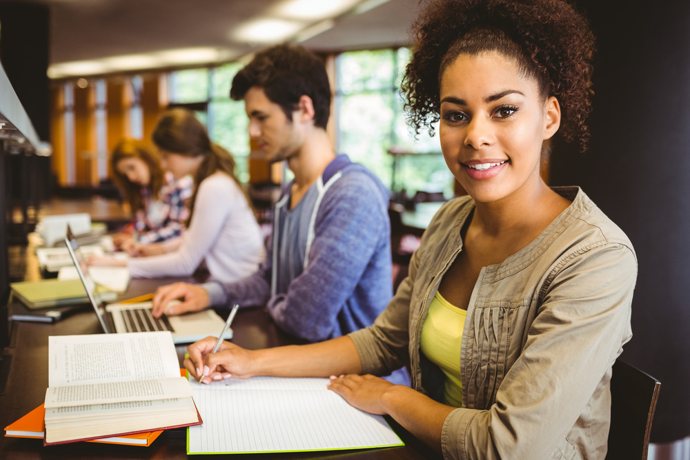 Student looking at camera while studying with classmates in library Student looking at camera while studying with classmates in library